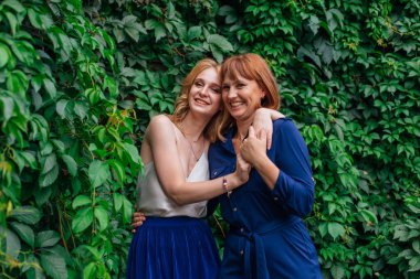 Güzel anne ve yetişkin kızı birbirine sarılıyor ve bir yaz günü dışarıda yeşil bir arka planda gülüyorlar.