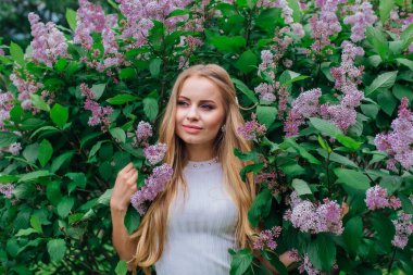 Güzel beyaz elbise giyen büyüleyici bir sarışın kadının bahar portresi çiçek açan mor leylak çalı yanında duran.