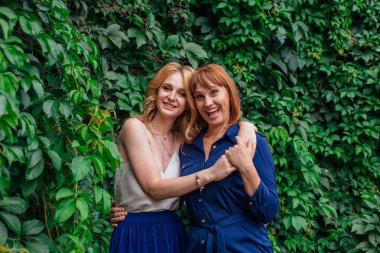 Güzel anne ve yetişkin kızı birbirine sarılıyor ve bir yaz günü dışarıda yeşil bir arka planda gülüyorlar.