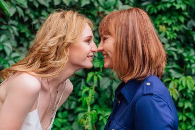 Güzel anne ve yetişkin kızı bir yaz günü yeşil bir arka planda burun buruna duruyorlar.