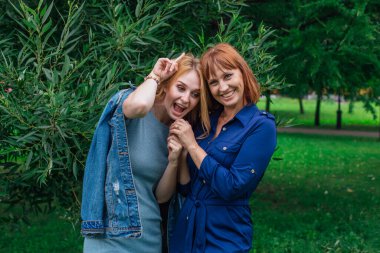 Güzel anne ve yetişkin kızı birbirine sarılıyor ve bir yaz günü dışarıda yeşil bir arka planda gülüyorlar.