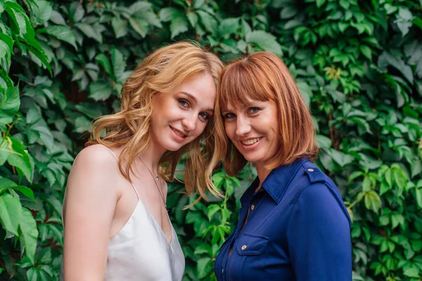 Güzel anne ve yetişkin kızı bir yaz günü yeşil bir arka planda kafa kafaya duruyorlar.