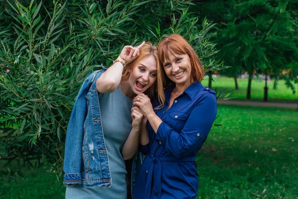 Güzel anne ve yetişkin kızı birbirine sarılıyor ve bir yaz günü dışarıda yeşil bir arka planda gülüyorlar.
