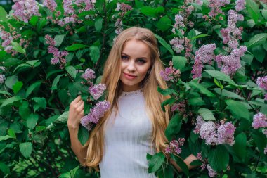 Güzel beyaz elbise giyen büyüleyici bir sarışın kadının bahar portresi çiçek açan mor leylak çalı yanında duran.