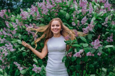 Güzel beyaz elbiseler giyen çekici bir sarışının bahar portresi yapraklarında saçı olan mor leylak çalılarının yanında duruyor.