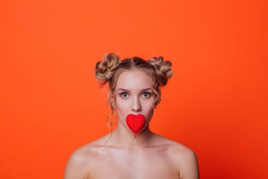 Stüdyoda turuncu arka plandaki kız. Sarışın kız, iki saç düğümlü. Kırmızı kalbi dudaklarına yakın tutuyor..