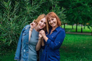 Güzel anne ve yetişkin kızı birbirine sarılıyor ve bir yaz günü dışarıda yeşil bir arka planda gülüyorlar.
