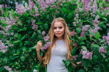 Güzel beyaz elbise giyen büyüleyici bir sarışın kadının bahar portresi çiçek açan mor leylak çalı yanında duran.