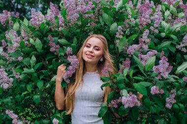 Güzel beyaz elbise giyen büyüleyici bir sarışın kadının bahar portresi çiçek açan mor leylak çalı yanında duran.