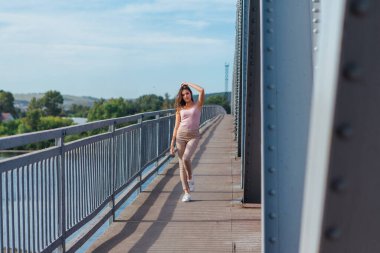 Gün batımında nehrin üzerindeki eski paslı nakliye köprüsünde poz veren genç ve güzel bir kadının ılık yaz portresi..