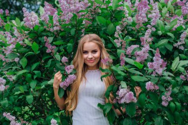 Güzel beyaz elbise giyen büyüleyici bir sarışın kadının bahar portresi çiçek açan mor leylak çalı yanında duran.