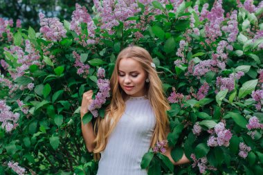 Güzel beyaz elbise giyen büyüleyici bir sarışın kadının bahar portresi çiçek açan mor leylak çalı yanında duran.