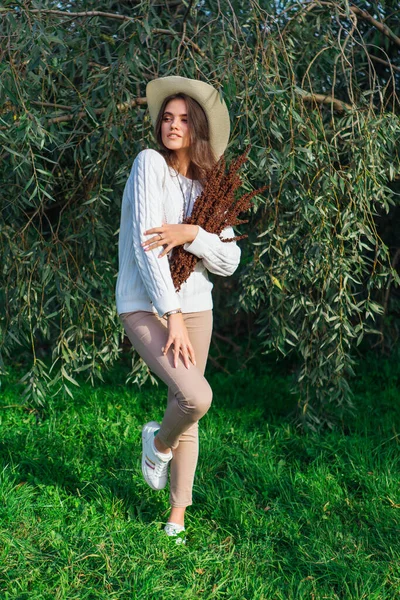 Beyaz kazak, kot pantolon ve kovboy hasır şapkası giymiş, elinde kuru yapraklarla ağaç brançlarının arkasında duran mutlu esmer kız.