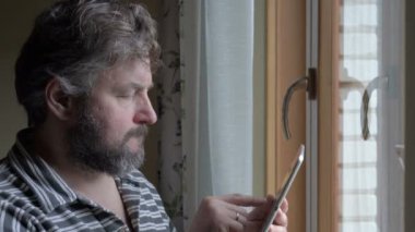 Erkek işadamı kahve içiyor ve pencere kenarında tablet üzerinde çalışıyor. İşinden memnun.