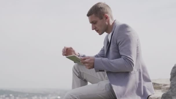 Young dreamed businessman with tablet. He sits on a hump and works on a tablet. — 비디오