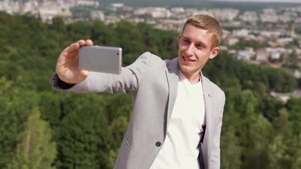 Бизнесмен стоит на холме и делает селфи на фоне города — стоковое видео