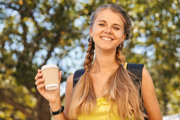 Красивая молодая студентка пьет кофе по дороге в колледж или университет. Мода, одетая для школы, с книгами и блокнотом в солнечных очках . — стоковое фото