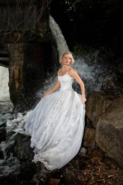 年轻的金发女人穿着白色婚纱在瀑布附近 — 图库照片