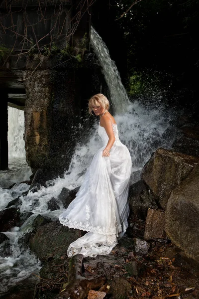 年轻的金发女人穿着白色婚纱在瀑布附近 — 图库照片