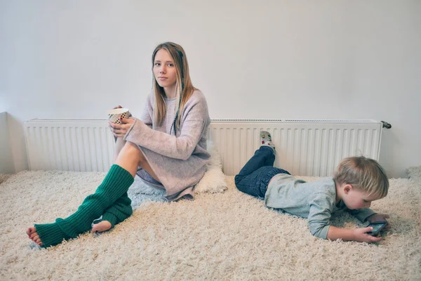 Blonde young mother sitting on the sofa with a cap in green socks and baby lying next played