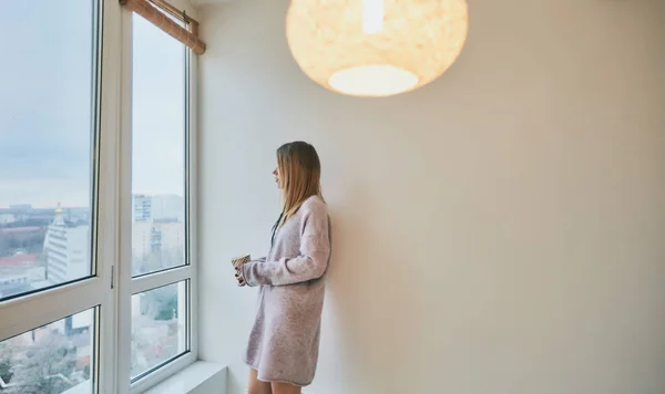 Hermosa chica en calcetines verdes y suéter largo de color púrpura pálido con una taza de té mirando a la ventana, el hogar, la comodidad, la felicidad —  Fotos de Stock