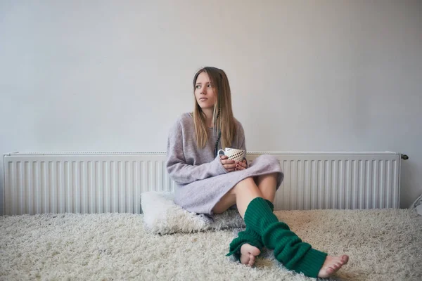 Hermosa chica en calcetines verdes y suéter largo de color púrpura pálido con una taza de té sentado en una cama cerca de la ventana, el hogar, la comodidad, la felicidad —  Fotos de Stock