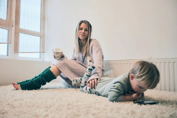 Blonde young mother sitting on the sofa with a cap in green socks and baby lying next played