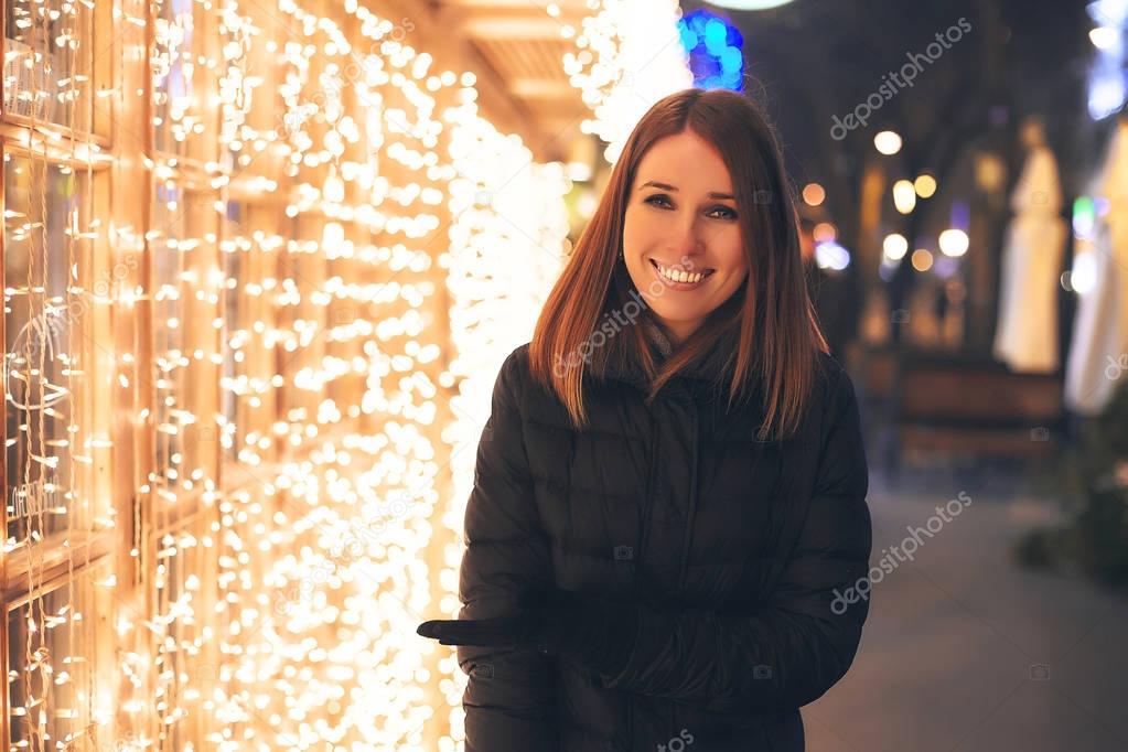 beautiful girl stands and smiles in the street next to the coffee shop, which is decorated with beautiful garlands, Christmas parties, New Year's holiday