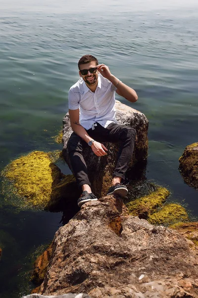 Jovem homem em óculos de sol sentado em rochas — Fotografia de Stock