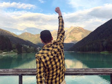 Gündüz vakti ormanla kaplı dağların arasında gölün kıyısında duran yükseltilmiş kollu bir erkek gezginin arka görüntüsü. 