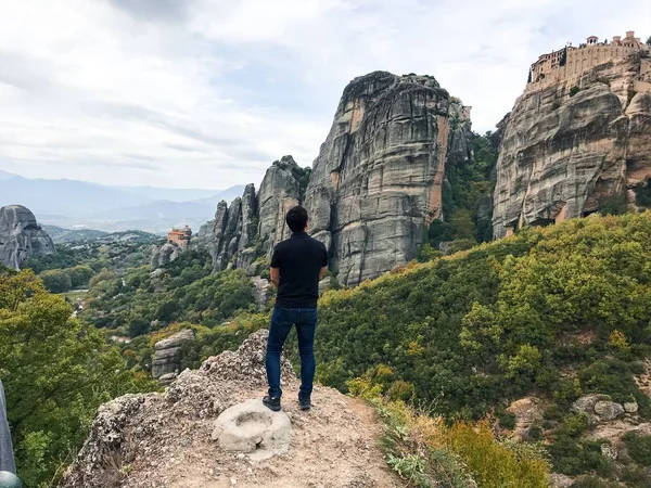 back view of man standing among mountains covered with forest near building on top of rock at daytime