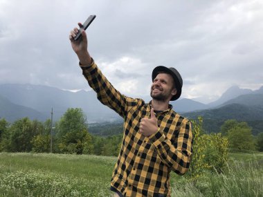 Gündelik giysiler içinde yakışıklı bir adam akıllı telefondan selfie çekiyor ve yaz günü dağların arasında işaret veriyor.  