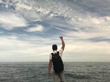 Güneş gözlüklü, sırt çantalı, kayalık deniz kıyısında yaz günü poz veren adamın arka görüntüsü.