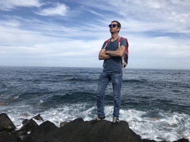 Güneş gözlüklü ve sırt çantalı bir adam yaz günü denizin kayalık kıyılarında poz veriyor.