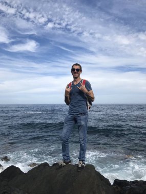 Güneş gözlüklü ve sırt çantalı bir adam yaz günü denizin kayalık kıyılarında poz veriyor.