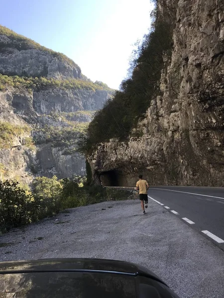 back view of man running on empty road among high rocky mountains covered with forest at daytime