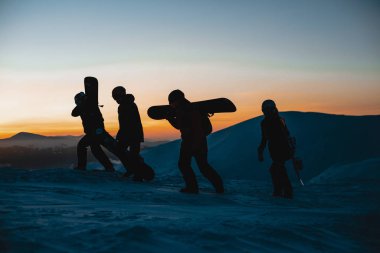 Kışın snowboardcular ellerinde snowboardla kış günbatımında tepeye tırmanıyorlar.   
