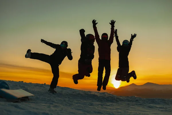 silhouettes of friends in winter clothes jumping together on hill in winter mountains during sunset