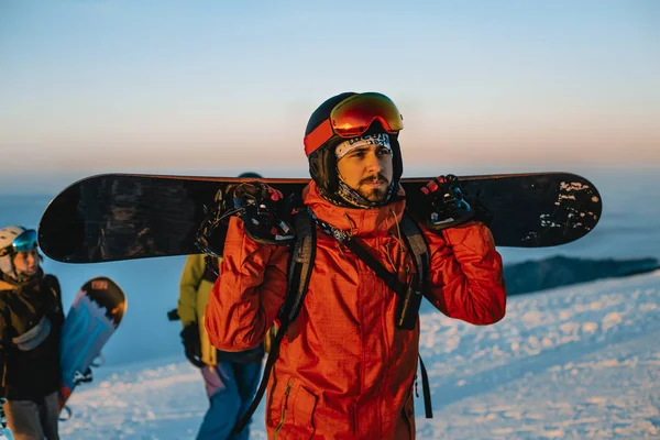 man holding snowboard and walking with friends in winter mountains