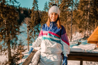 beautiful woman in warm clothes leaning on railing while standing snow-covered forest at sunny winter day 