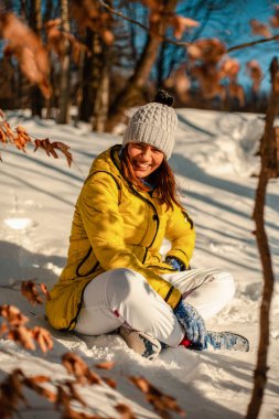 smiling woman in warm clothes looking and camera and sitting on snow outside at sunny winter day 