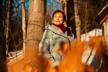 beautiful adult woman in warm clothes leaning on tree in winter forest at sunny day  
