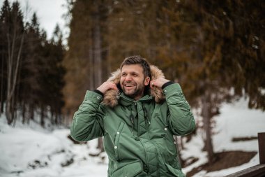 happy bearded man in warm coat sitting on bridge railing in snow-covered winter forest 