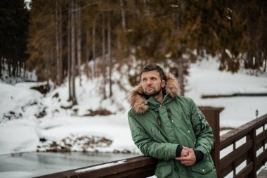 happy bearded man in warm coat standing on wooden bridge in snow-covered winter forest 
