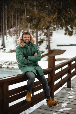 happy bearded man in warm coat sitting with crossed arms on bridge railing in snow-covered winter forest 