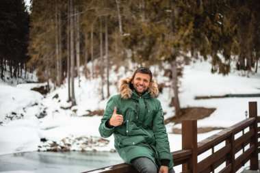 happy bearded man in warm coat showing thumb up and sitting on bridge railing in snow-covered winter forest 