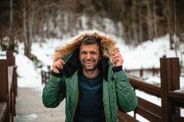 happy bearded man in warm coat standing on wooden bridge in snow-covered winter forest 
