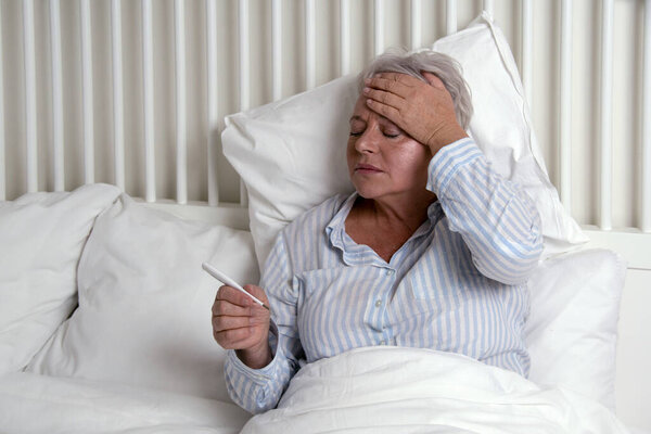 Старшая женщина, лежащая в постели, страдает от лихорадки, проверяющей температуру на термометре. Концепция опасности коронавируса для пожилых людей
