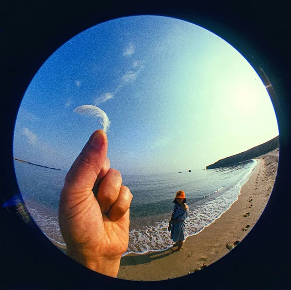 概念照片 在海滨形成一个圆形的球体 在前面 一只手拿着蓬松的白羽 沙子里有个戴帽子的女孩 用的是鱼眼灯 — 图库照片