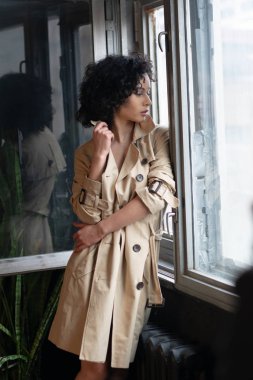 Genç güzel kız pencerenin yanında yağmurlukla duruyor.